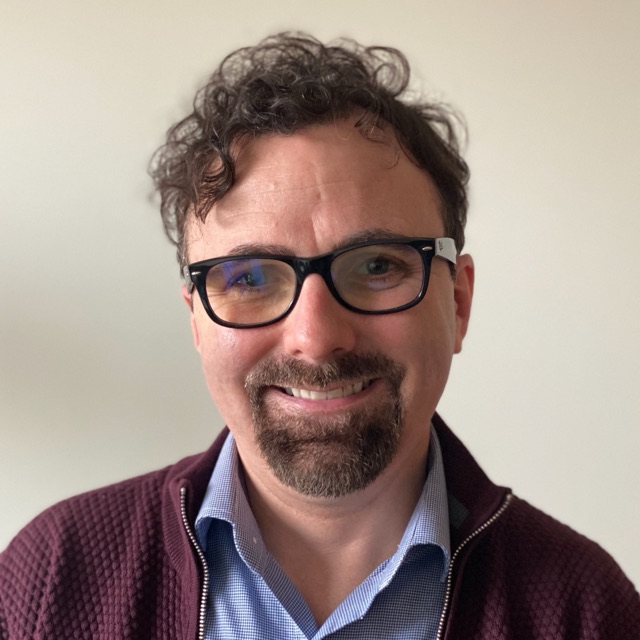 Adam Witwer, smiling, in glasses and a maroon cardigan